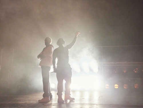 12 Two silhouetted figures stand with back-to-back with bright lights blaring behind them. One has their arms folded, the other has one by their side holding a mic, the other raised in the air, pointing upwards. Both face out away from the lights.